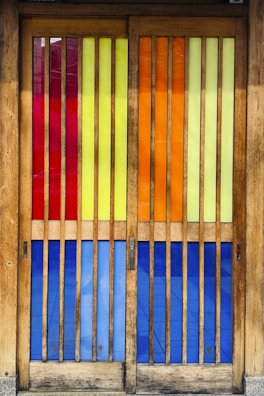 A set of wooden doors featuring vertical slats of colorful stained glass panels. The glass sections include rich hues of red, yellow, orange, and blue, creating a vibrant and eye-catching effect. The wooden frame has a natural finish, complementing the bright colors and adding warmth to the visual composition.
