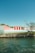 Several storage units with vibrant red doors are situated along a rocky shoreline, with clear blue skies overhead and some sparse trees nearby.