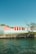 Several storage units with vibrant red doors are situated along a rocky shoreline, with clear blue skies overhead and some sparse trees nearby.