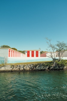 Several storage units with vibrant red doors are situated along a rocky shoreline, with clear blue skies overhead and some sparse trees nearby.