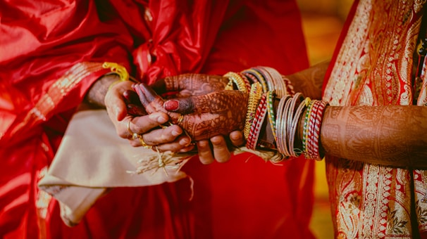 Two hands are clasped together, adorned with intricate henna designs. One hand wears numerous bangles and a traditional red and gold saree sleeve is visible. This appears to be a scene from a cultural or wedding ceremony.