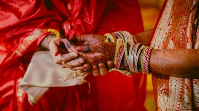 Two hands are clasped together, adorned with intricate henna designs. One hand wears numerous bangles and a traditional red and gold saree sleeve is visible. This appears to be a scene from a cultural or wedding ceremony.