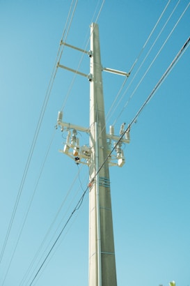 A tall electricity pole stands against a clear blue sky, with multiple lines of cables stretching outwards. The pole is equipped with insulators and other electrical components.