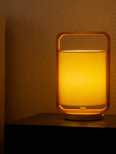 A modern lamp with a wooden frame emits a warm yellow light, creating a cozy ambiance. The lamp is placed on a dark surface against a textured, patterned wall.