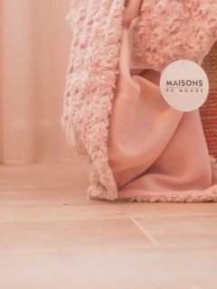 A soft textured pink fabric drapes loosely on a wooden floor, accompanied by a label bearing the name 'Maisons du Monde'. The fabric appears to have a plush, cozy texture.