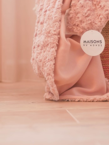 A soft textured pink fabric drapes loosely on a wooden floor, accompanied by a label bearing the name 'Maisons du Monde'. The fabric appears to have a plush, cozy texture.