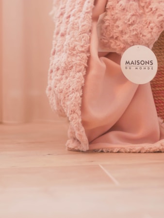 A soft textured pink fabric drapes loosely on a wooden floor, accompanied by a label bearing the name 'Maisons du Monde'. The fabric appears to have a plush, cozy texture.