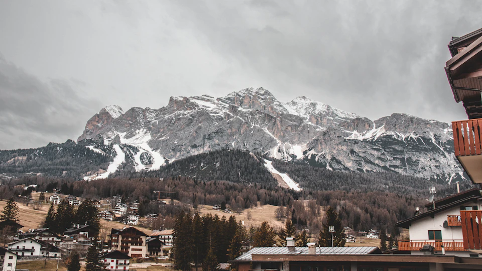 The image depicts a mountainous landscape with a large, snow-capped mountain range in the background and a smaller mountain range in the foreground. The sky is overcast, casting a soft light on the scene below. In the foreground, there are several buildings nestled among the trees, including a white building with a red roof that stands out against the greenery.