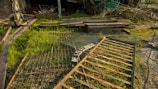 A grassy area with scattered debris and scrap metal pieces, including a weathered gate lying on the ground. There are wooden planks and branches piled in the background against a dilapidated structure.