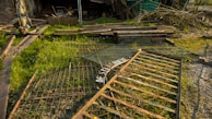A grassy area with scattered debris and scrap metal pieces, including a weathered gate lying on the ground. There are wooden planks and branches piled in the background against a dilapidated structure.