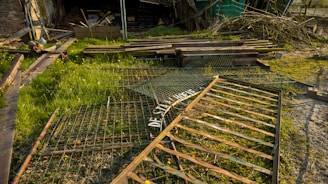 A grassy area with scattered debris and scrap metal pieces, including a weathered gate lying on the ground. There are wooden planks and branches piled in the background against a dilapidated structure.