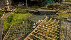 A grassy area with scattered debris and scrap metal pieces, including a weathered gate lying on the ground. There are wooden planks and branches piled in the background against a dilapidated structure.