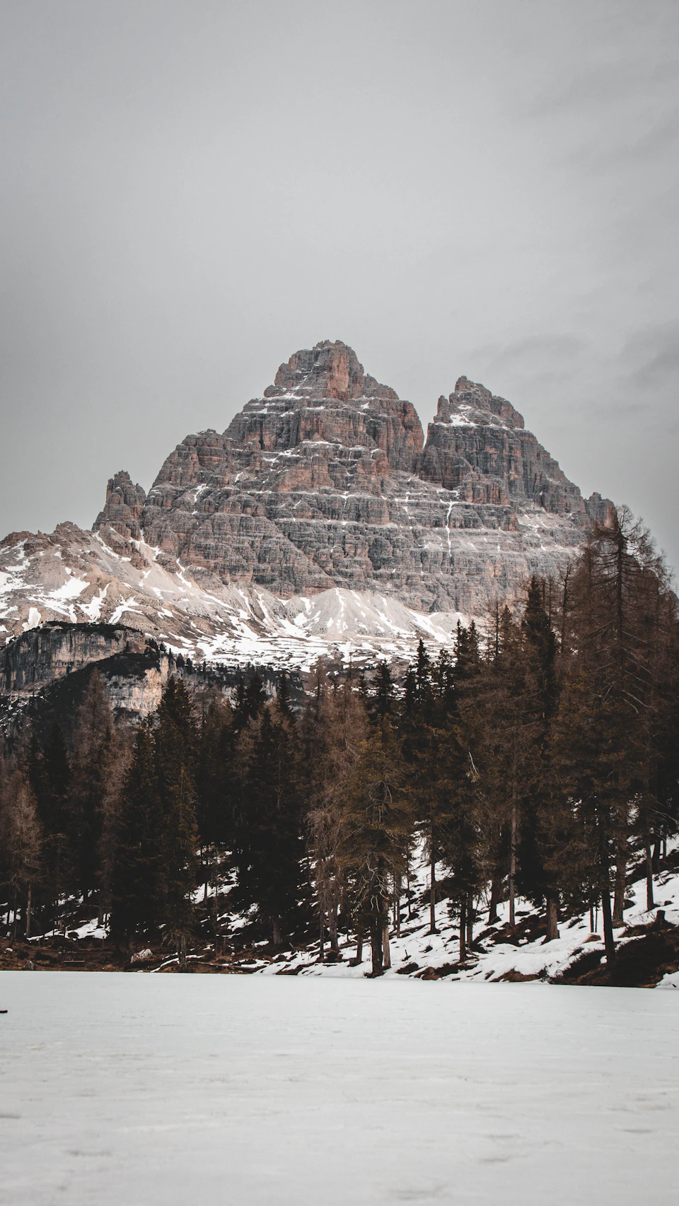 The image depicts a majestic mountain range with snow-capped peaks and a deep blue sky above. The mountains, covered in white snow, rise high into the sky, creating a stunning backdrop for the scene. In the foreground, there is a frozen lake surrounded by trees on both sides, adding to the serene atmosphere of the image.