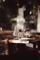 A wooden dining table is elegantly set with multiple wine glasses, plates, and cutlery in a dimly lit restaurant setting. The background is softly blurred, enhancing the intimate and cozy atmosphere.
