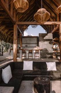 A rustic outdoor seating area featuring a black L-shaped couch with white pillows and a round wooden coffee table. In the background, there's a bar area with three wooden bar stools and a thatched roof. The structure includes several large, round, woven pendant lights hanging from the ceiling, with natural light streaming in.