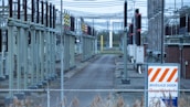 A fenced-off area containing what appears to be electrical transformers and various utility poles with numerous wires overhead. The setup is organized in rows with colored indicators on equipment. A warning sign with an orange and white pattern is visible in the foreground.