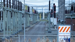 A fenced-off area containing what appears to be electrical transformers and various utility poles with numerous wires overhead. The setup is organized in rows with colored indicators on equipment. A warning sign with an orange and white pattern is visible in the foreground.
