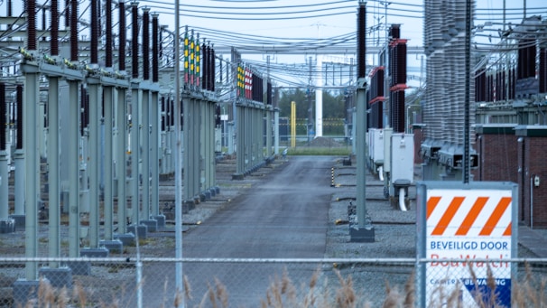 A fenced-off area containing what appears to be electrical transformers and various utility poles with numerous wires overhead. The setup is organized in rows with colored indicators on equipment. A warning sign with an orange and white pattern is visible in the foreground.