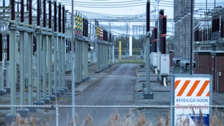 A fenced-off area containing what appears to be electrical transformers and various utility poles with numerous wires overhead. The setup is organized in rows with colored indicators on equipment. A warning sign with an orange and white pattern is visible in the foreground.