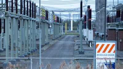 A fenced-off area containing what appears to be electrical transformers and various utility poles with numerous wires overhead. The setup is organized in rows with colored indicators on equipment. A warning sign with an orange and white pattern is visible in the foreground.