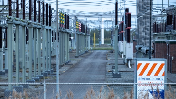 A fenced-off area containing what appears to be electrical transformers and various utility poles with numerous wires overhead. The setup is organized in rows with colored indicators on equipment. A warning sign with an orange and white pattern is visible in the foreground.