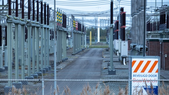 A fenced-off area containing what appears to be electrical transformers and various utility poles with numerous wires overhead. The setup is organized in rows with colored indicators on equipment. A warning sign with an orange and white pattern is visible in the foreground.
