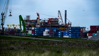 An industrial area with multiple shipping containers stacked high, cranes, and other construction machinery. The machinery includes a prominent green crane and a large yellow structure. The sky appears overcast, adding a moody atmosphere to the scene.