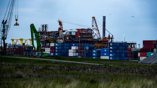 An industrial area with multiple shipping containers stacked high, cranes, and other construction machinery. The machinery includes a prominent green crane and a large yellow structure. The sky appears overcast, adding a moody atmosphere to the scene.