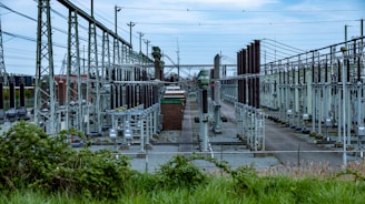 An extensive outdoor electrical substation features numerous transformers, circuit breakers, and metal structures interconnected by a grid of cables. The area is fenced, and the ground is covered in gravel. Overhead power lines are prominently visible along with high-voltage equipment.