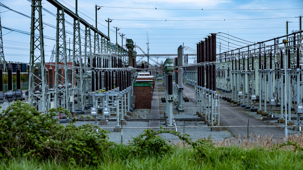 An extensive outdoor electrical substation features numerous transformers, circuit breakers, and metal structures interconnected by a grid of cables. The area is fenced, and the ground is covered in gravel. Overhead power lines are prominently visible along with high-voltage equipment.