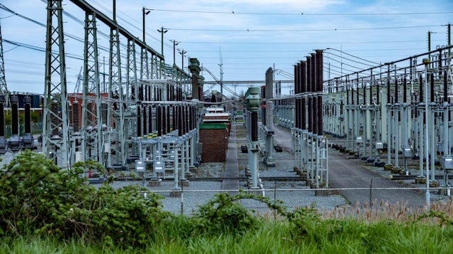 An extensive outdoor electrical substation features numerous transformers, circuit breakers, and metal structures interconnected by a grid of cables. The area is fenced, and the ground is covered in gravel. Overhead power lines are prominently visible along with high-voltage equipment.