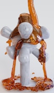 A viscous, amber-colored liquid flows over a geometrically shaped, white, wooden structure with cylindrical and spherical supports. The liquid cascades down and pools at the base of the structure, creating a glossy, reflective surface.