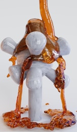 A viscous, amber-colored liquid flows over a geometrically shaped, white, wooden structure with cylindrical and spherical supports. The liquid cascades down and pools at the base of the structure, creating a glossy, reflective surface.