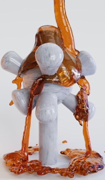 A viscous, amber-colored liquid flows over a geometrically shaped, white, wooden structure with cylindrical and spherical supports. The liquid cascades down and pools at the base of the structure, creating a glossy, reflective surface.