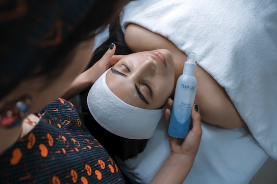 A person is lying down with a white headband, receiving a facial treatment. Another person is holding a skincare product labeled 'THAI AC' close to the face. The setting appears to be a salon or spa, indicated by the white towels and professional setup.