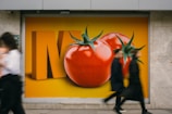 A large advertisement featuring oversized, vibrant tomatoes against an orange background is displayed on a wall. Two blurred figures walk past, suggesting motion and urban life.