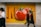 A large advertisement featuring oversized, vibrant tomatoes against an orange background is displayed on a wall. Two blurred figures walk past, suggesting motion and urban life.