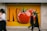 A large advertisement featuring oversized, vibrant tomatoes against an orange background is displayed on a wall. Two blurred figures walk past, suggesting motion and urban life.