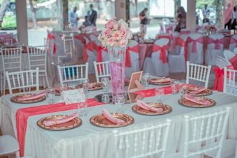 A beautifully decorated banquet table set for a formal event. The tables are adorned with pink and white floral arrangements in tall vases, gold-rimmed plates, pink napkins, and clear glassware. Chairs are elegantly covered with white fabric and tied with pink sashes. Other tables in the background echo the same decoration theme, contributing to a festive atmosphere.