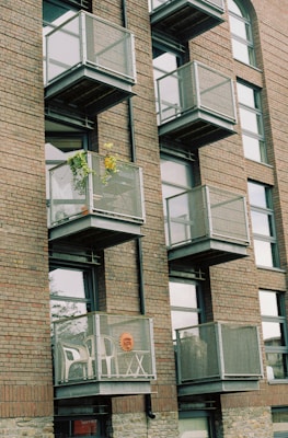Several modern apartment balconies attached to a brick building, featuring metal railings. One balcony contains a potted plant with green leaves and yellow flowers, while another has two white plastic chairs and a decorative item resembling a sun face.