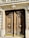 A pair of ornate wooden doors with intricate carvings and large circular brass handles are set within a stone façade. Above the doors, decorative stone molding and balustrades add to the architectural detail.