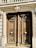 A pair of ornate wooden doors with intricate carvings and large circular brass handles are set within a stone façade. Above the doors, decorative stone molding and balustrades add to the architectural detail.