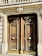 A pair of ornate wooden doors with intricate carvings and large circular brass handles are set within a stone façade. Above the doors, decorative stone molding and balustrades add to the architectural detail.