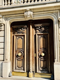 A pair of ornate wooden doors with intricate carvings and large circular brass handles are set within a stone façade. Above the doors, decorative stone molding and balustrades add to the architectural detail.