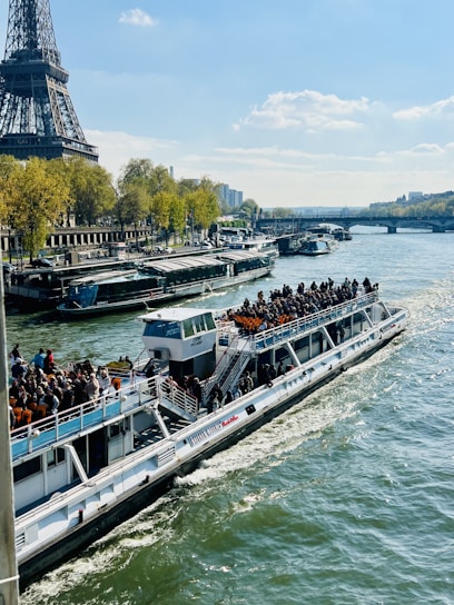 A riverboat filled with people cruises along the Seine River, with the Eiffel Tower prominently visible on the left side. The river is bustling with boats, and the opposite bank is lined with green trees and classic Parisian architecture. The sky is clear with a few clouds, indicating a sunny day.