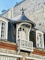 An ornate window with a small decorative balcony, framed by dark slate roofing and set against a textured stone and brick wall. The architectural style features elegant curves and detailed molding, adding a touch of classic elegance to the building facade.
