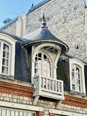 An ornate window with a small decorative balcony, framed by dark slate roofing and set against a textured stone and brick wall. The architectural style features elegant curves and detailed molding, adding a touch of classic elegance to the building facade.