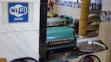 A vintage cash register is prominently positioned on a reflective surface next to a metal tray. A blue sign indicates the availability of WiFi. The setting includes ornate wall tiles and reflections of chairs and decor in the background, suggesting a cozy interior space.