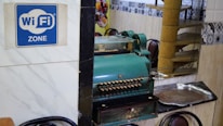 A vintage cash register is prominently positioned on a reflective surface next to a metal tray. A blue sign indicates the availability of WiFi. The setting includes ornate wall tiles and reflections of chairs and decor in the background, suggesting a cozy interior space.