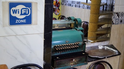 A vintage cash register is prominently positioned on a reflective surface next to a metal tray. A blue sign indicates the availability of WiFi. The setting includes ornate wall tiles and reflections of chairs and decor in the background, suggesting a cozy interior space.